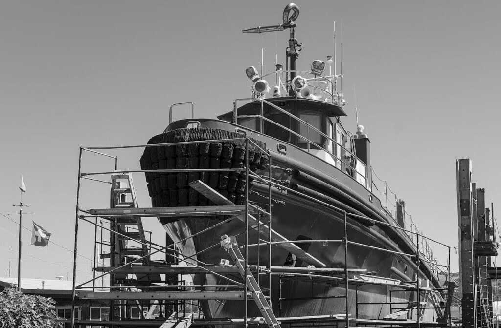 Monochrome image of ship undergoing repairs in a shipyard, showcasing industrial scaffolding.
