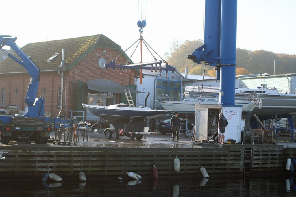 A boat being lifted for maintenance at an urban harbor with industrial buildings in the background.